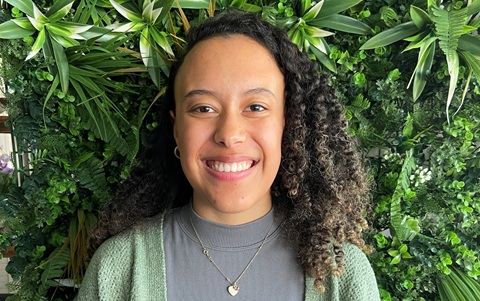Head and shoulders photo of a young woman smiling. She is stood in front of a green leafy background.