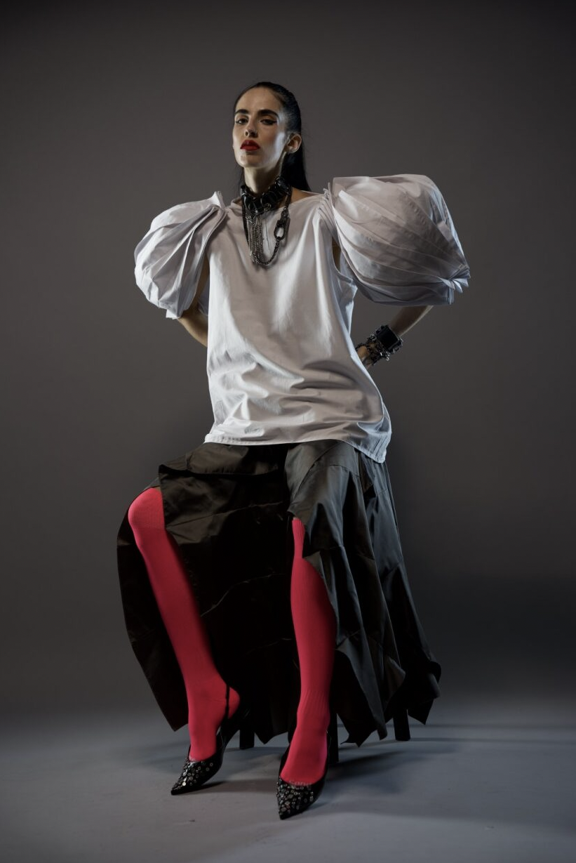 A person sits against a dark background wearing a white top with voluminous sleeves, a black layered skirt, bright pink tights, and black pointed shoes. They are accessorized with a necklace and bracelets.
