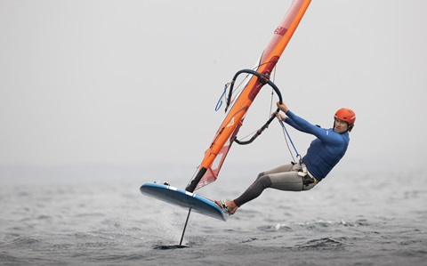 A man in a blue top and orange helmet windsurfing. The board is lifted out of the water by the foil beneath. 