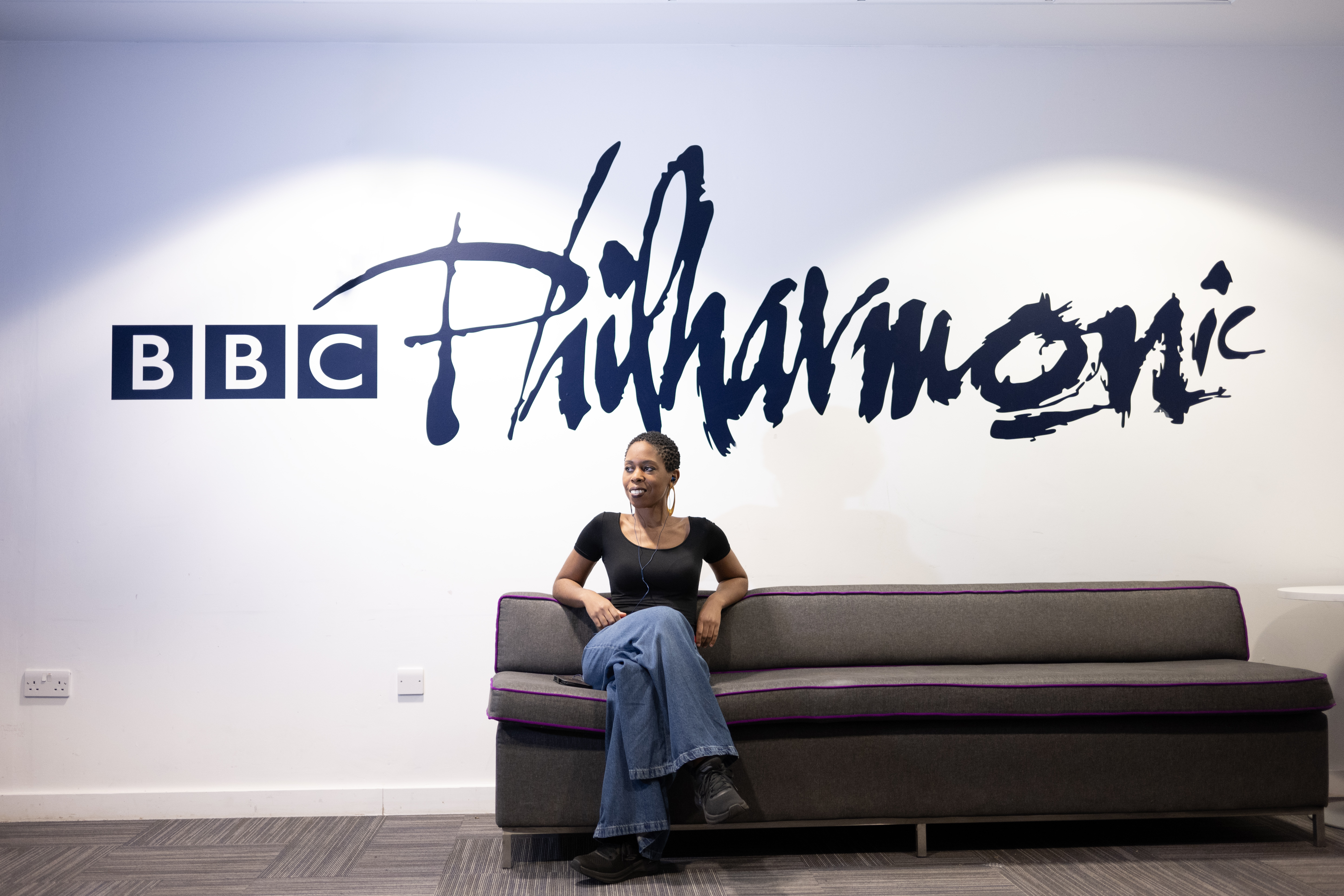A woman wearing a black top and blue jeans sits on a gray couch with purple trim, smiling and looking to the side. Behind her, the words 
