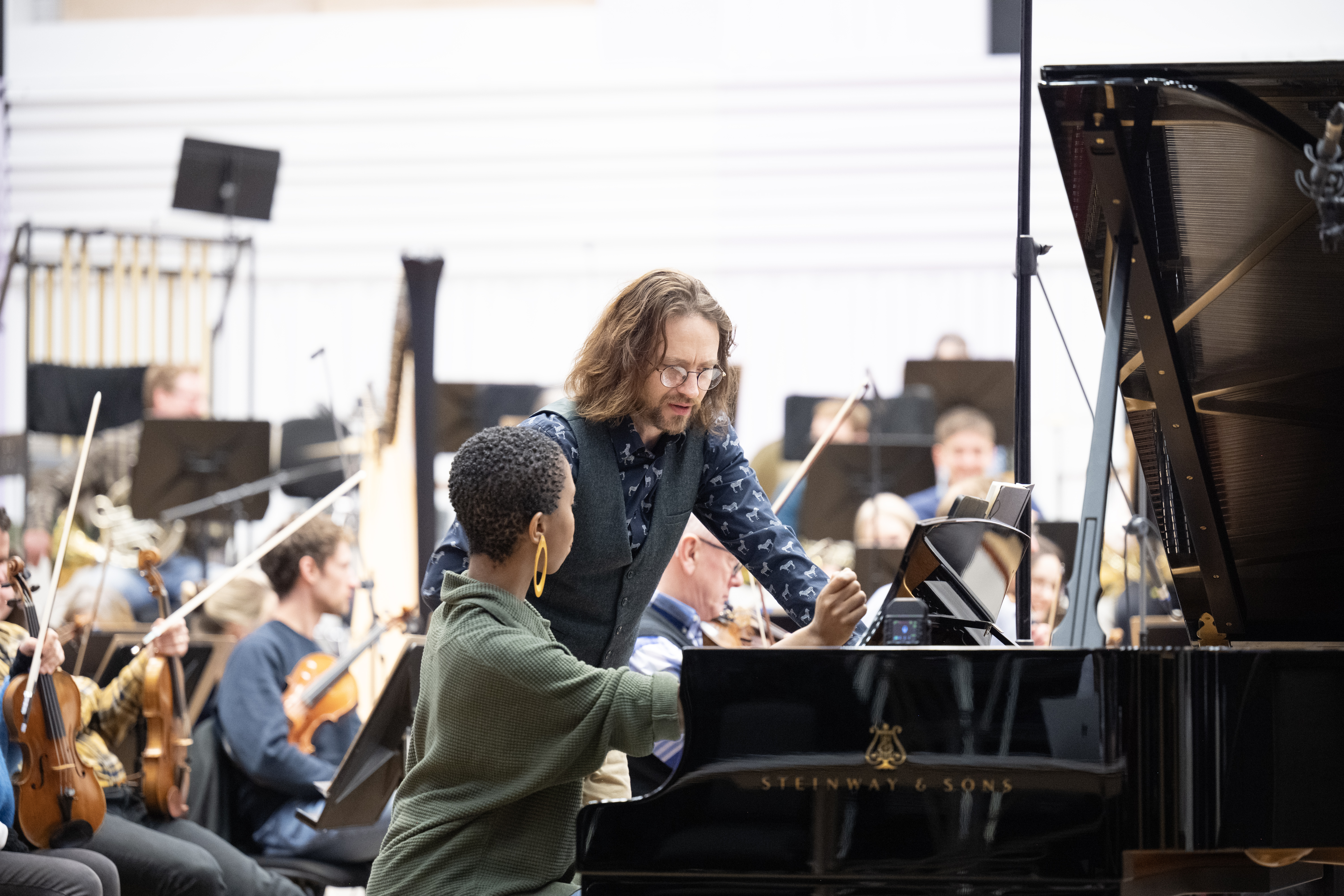 **Alt text:** A pianist in a green sweater and yellow earrings sits at a Steinway & Sons grand piano, engaged in conversation with a conductor who has shoulder-length hair and glasses. Behind them, an orchestra of musicians with string instruments is visible, focused on their performance.