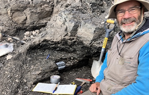 Bearded man wearing glasses, sun hat, beige gilet and blue long sleeved top sits on a rockside next to equipment including a spade, notebook and pens and a hammer