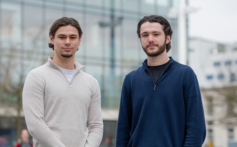 Two young men standing outside with a large glass-fronted building in the blurred background. The man on the left is wearing a cream coloured long sleeved top, and the man on the right is wearing a blue long sleeved top.