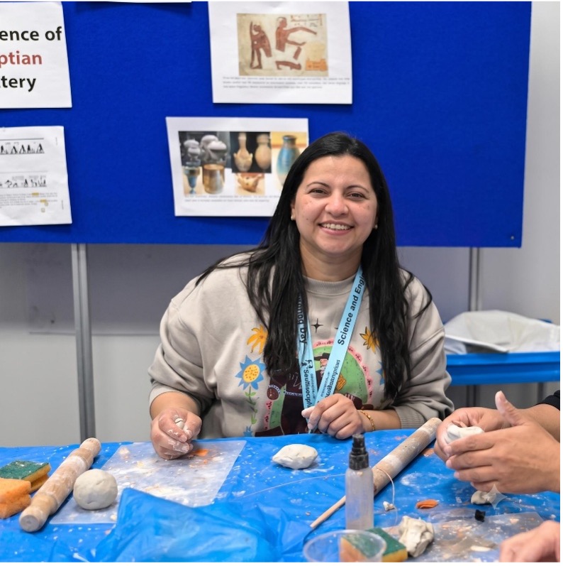 A person sits at a table shaping clay during a hands-on pottery activity, with rolling pins, clay pieces, and craft materials visible on the work surface. Behind the table, a display board features images and information related to ancient pottery and artefacts.