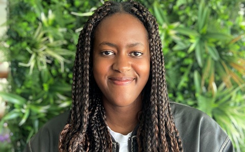 Head and shoulders photo of a young woman smiling at the camera. She is stood in front of a green leafy background
