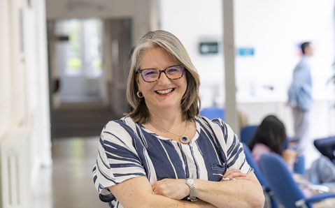 A woman with shoulder length fair har and glasses, wearing a blue and white striped short sleeved top, standing and smiling with her arms crossed.