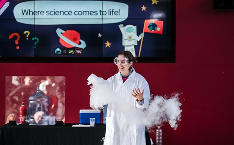 Woman in white lab coat holding a container of dry ice in front of a crowd. She’s standing in front of a red wall and a screen with the words ’where science comes to life’