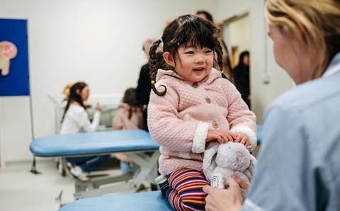 A young girl wearing a pink jacket and stripey trousers sitting on a hospital bed holding a fluffy toy bunny, talking to a nurse in uniform
