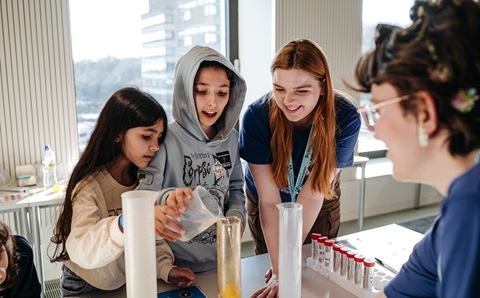 Two children pouring liquid from a jug into a tall glass tube, with two adults watching