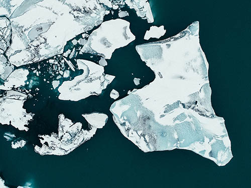 Aerial view of large white and pale blue sea ice floes floating in dark teal ocean water, with smaller fragments of broken ice scattered between them.