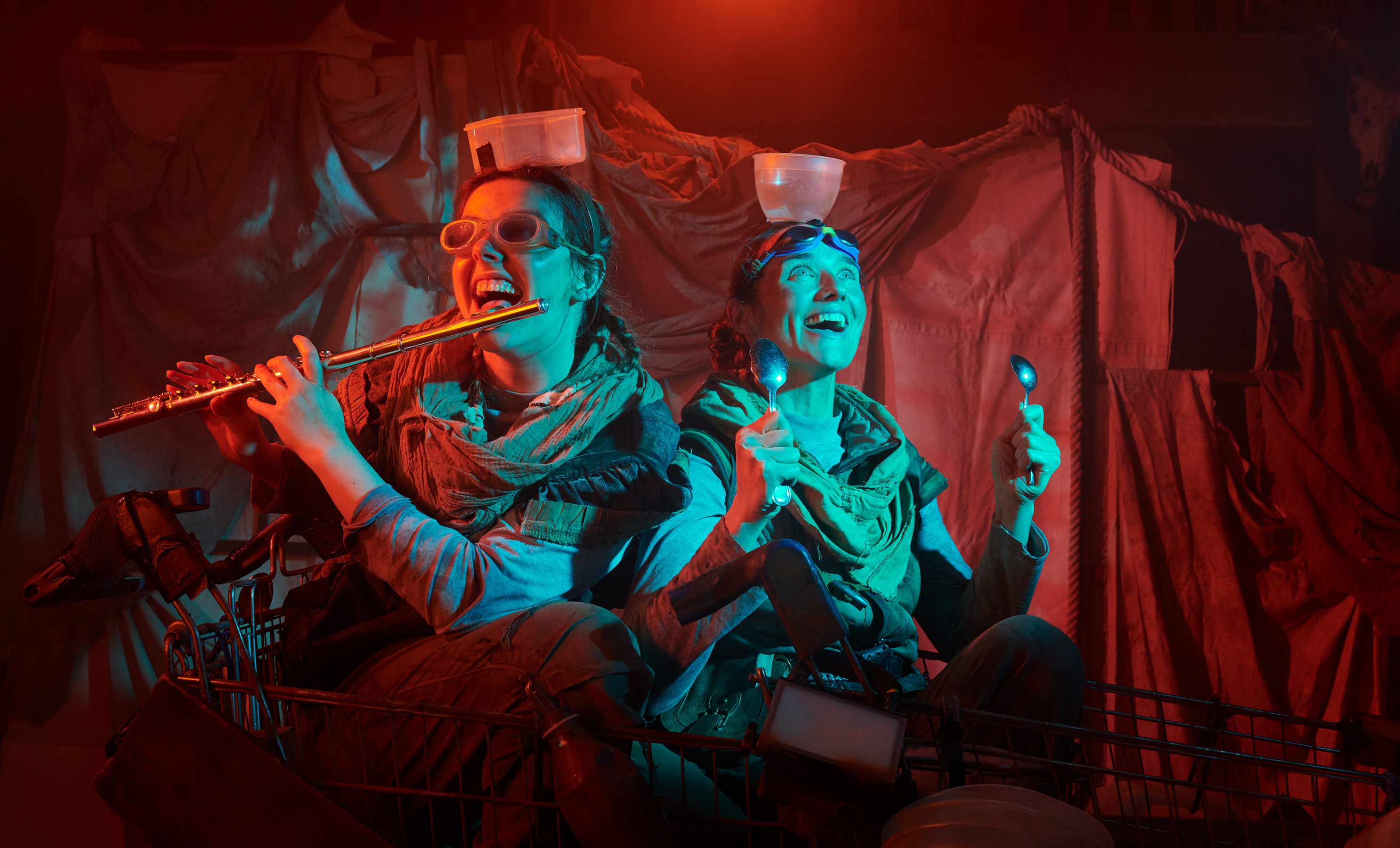 Two women smiling with props including a flute, goggles, spoons, and pots attached to the tops of their heads