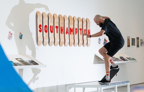 A man on a skateboard doing a jump, indoors in front of a white wall with skateboards hanging up, each one with a letter on spelling Southampton