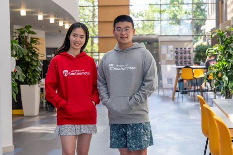 A student wearing a red hoodie standing next to a student wearing a grey hoodie. They are inside a building and both students have their hands in the front pocket.