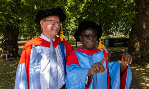 A man and a woman wearing red and blue graduation robes and black hats smiling, looking at a mobile phone and taking a selfie