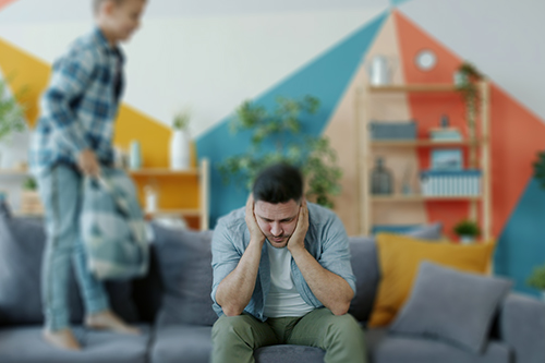 A stressed man sits on a sofa with his head in his hands while a young child jumps on the couch beside him in a colourful living room.
