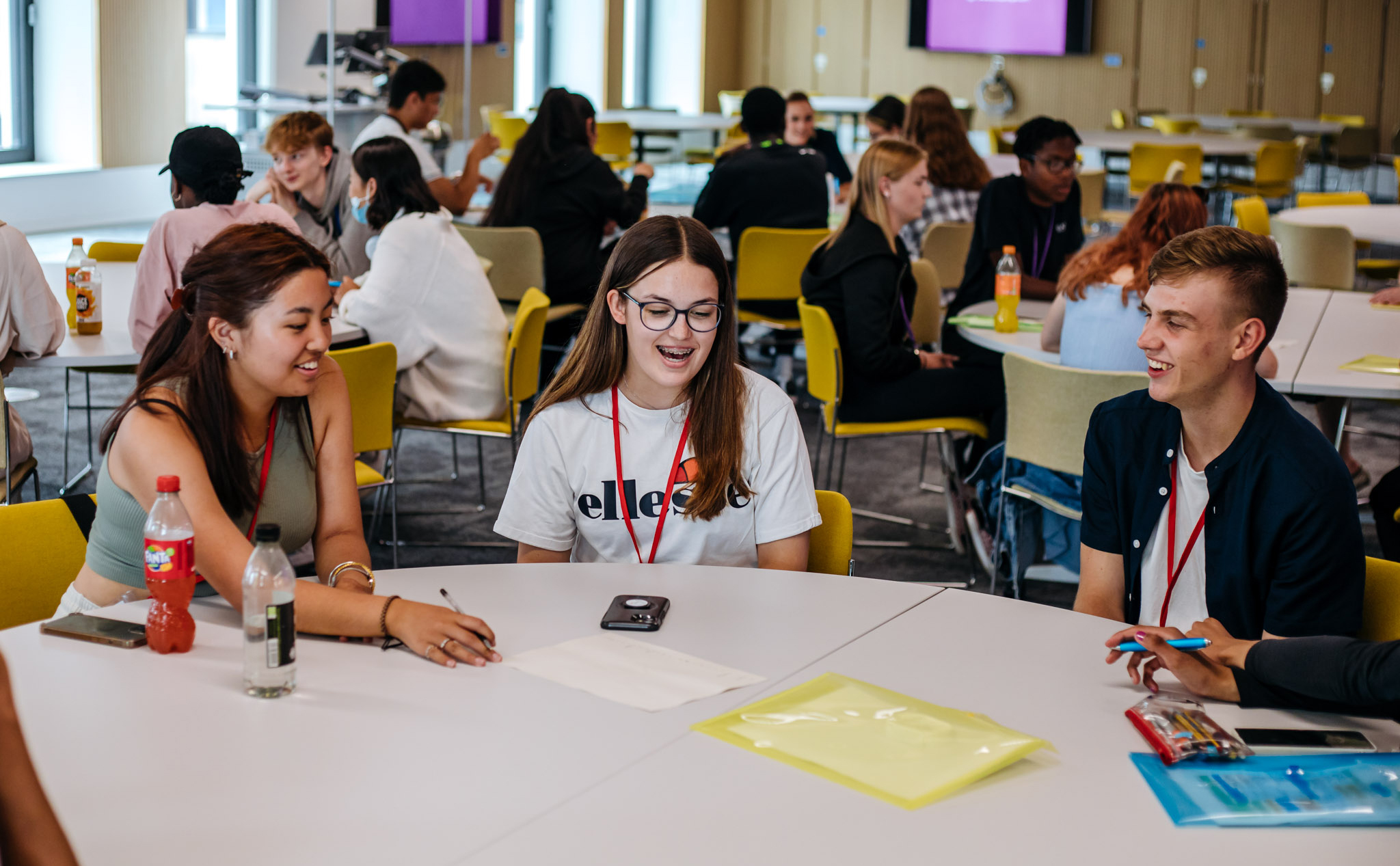Three young people, two female and one male, sat round a desk in a classroom environment smiling and studying.