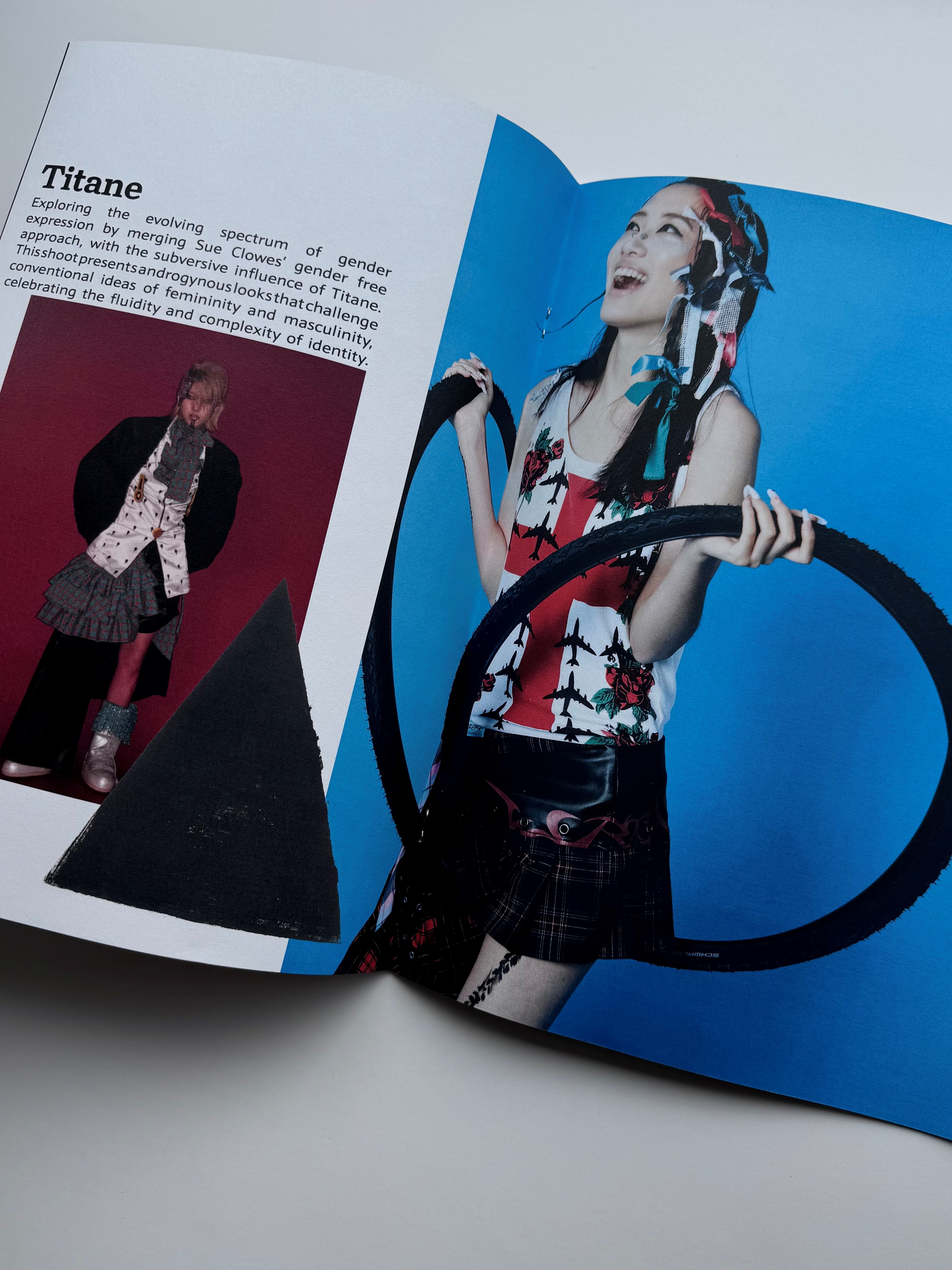A magazine is open on a double-page spread of a fashion photo shoot. Text on one page is titled ’Titane’ and there is an image of a model wearing an oversized jacket, shirt, scarf and ra-ra skirt. On the opposite page, a model wears a white vest emblazoned with a red cross and aircraft silhouettes with a tartan skirt. Their hair is decorated with red, white and blue bows and they are holding bicycle tyres. 