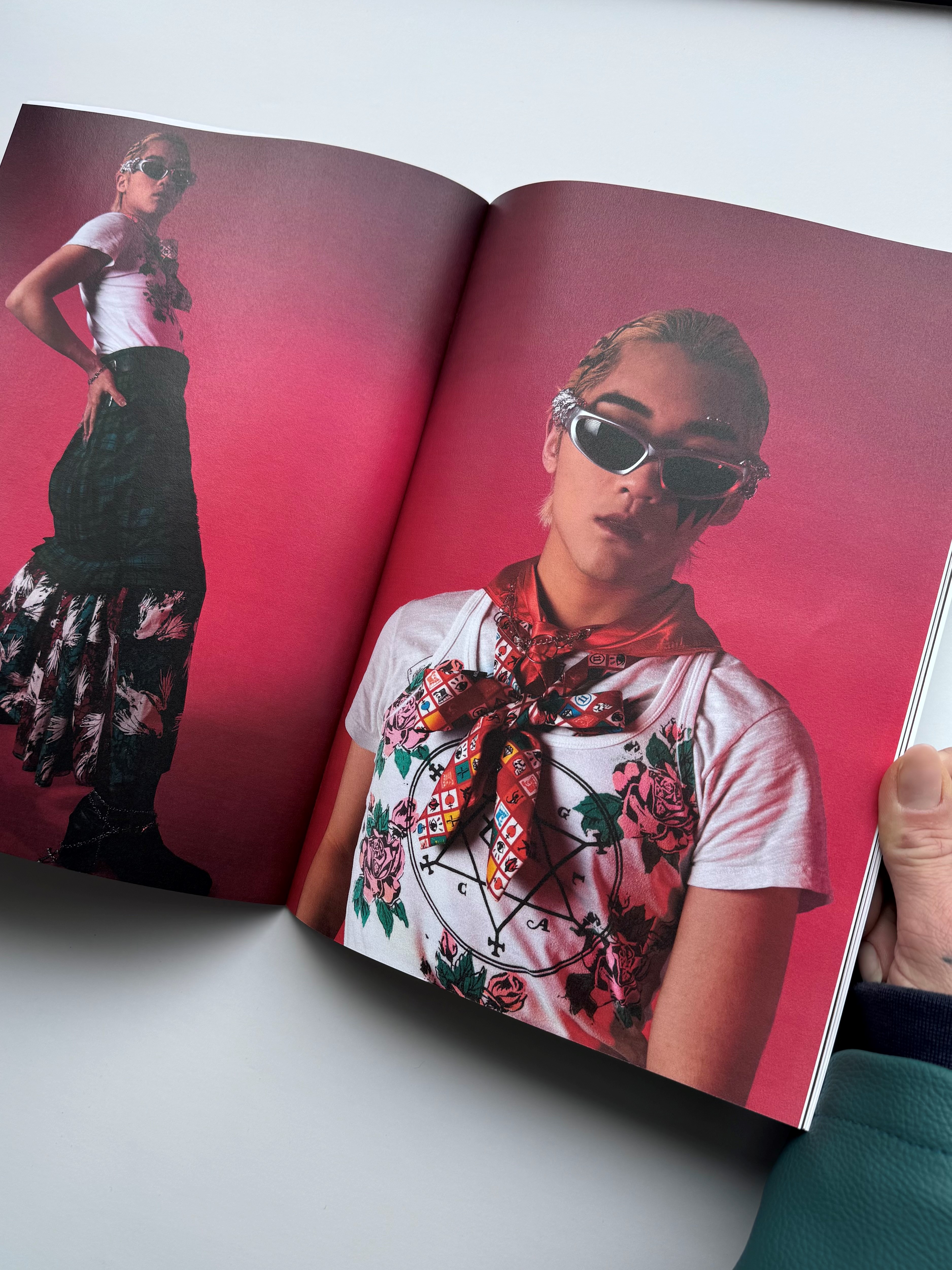 A magazine is open on a double-page spread of a fashion photo shoot. The model wears a white t-shirt with a pentagram and flowers on, and a tiered long skirt. The outfit is completed with a red print neckerchief and sporty silver sunglasses