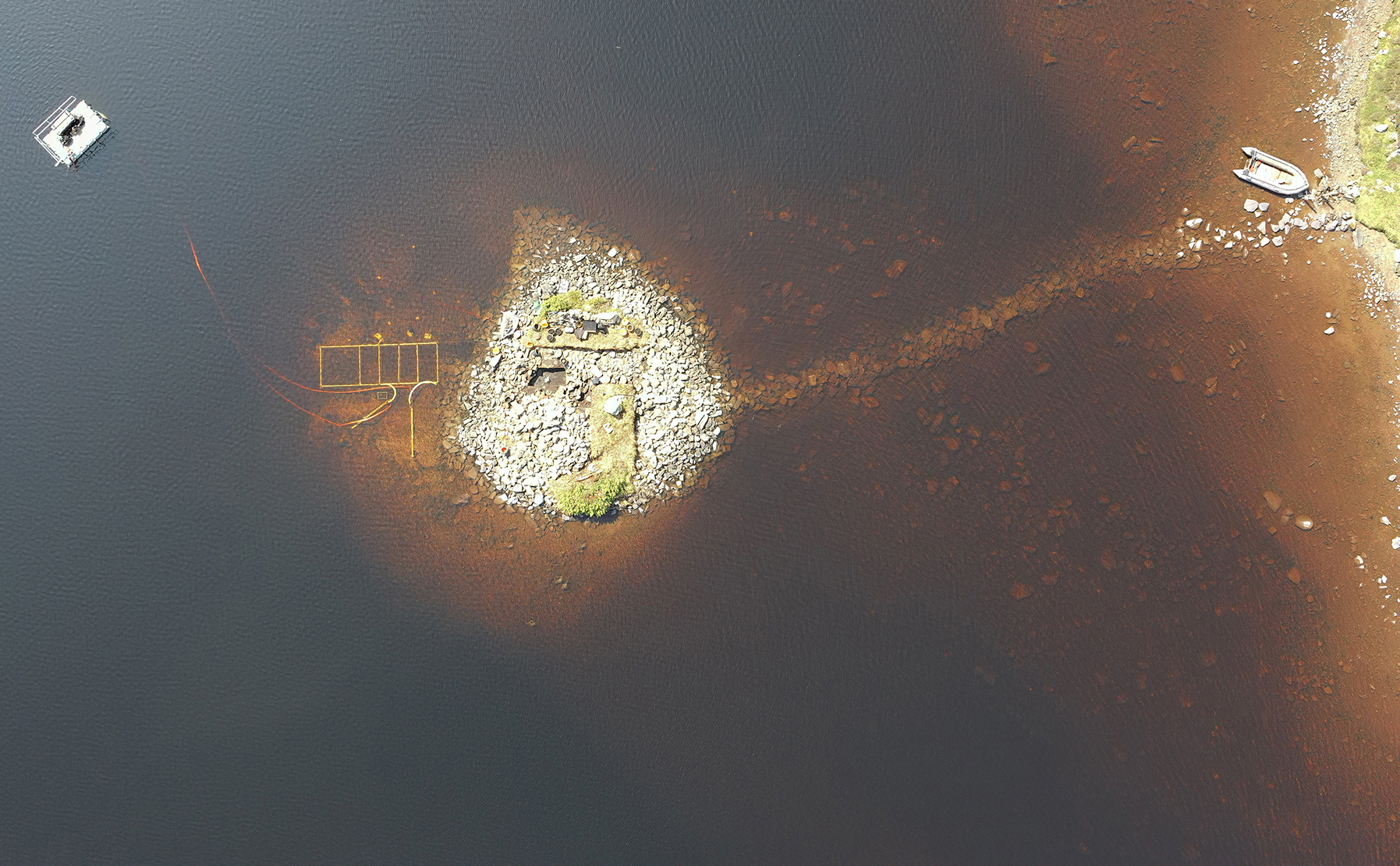 An aerial view of a small stony island in a lake with an inflatable boat nearby and a floating platform connected to scanning equipment on the island.
