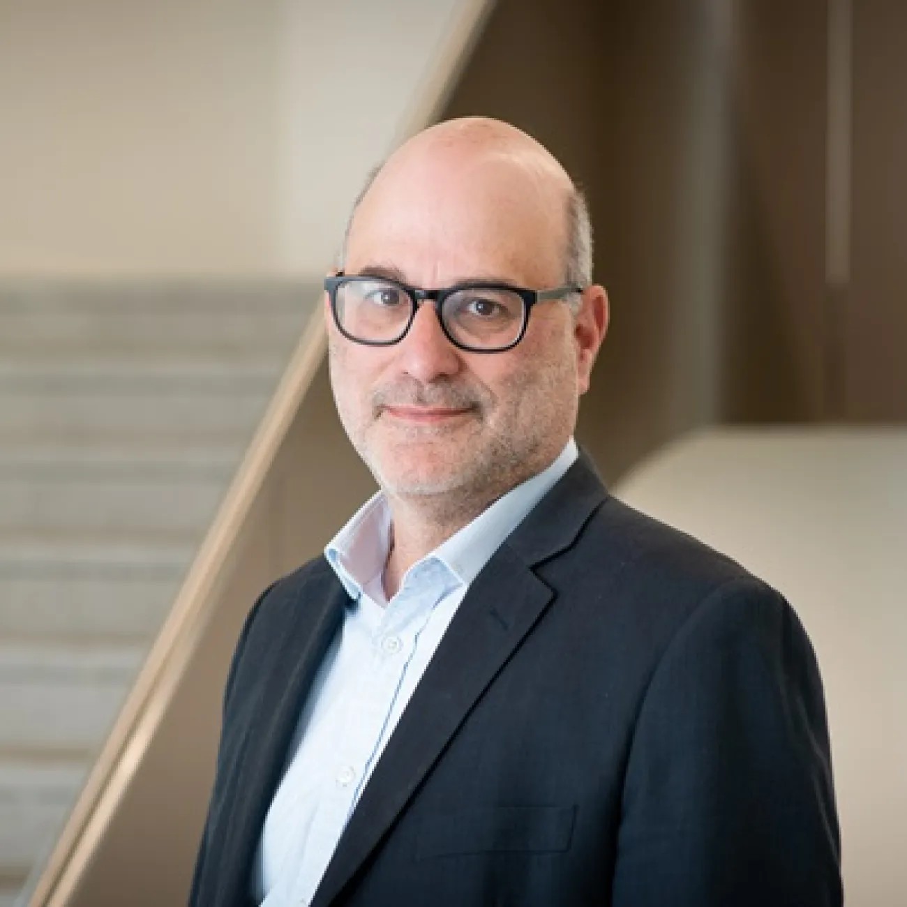 A person wearing glasses and a dark suit jacket stands indoors in front of a staircase and smooth architectural surfaces. Soft lighting highlights the individual against the neutral background.