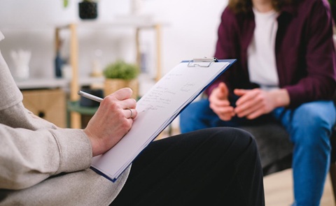 Close up of a woman’s hands taking notes using a pen and clipboard. Out of focus in the background is a person sitting with their hands in their lap.