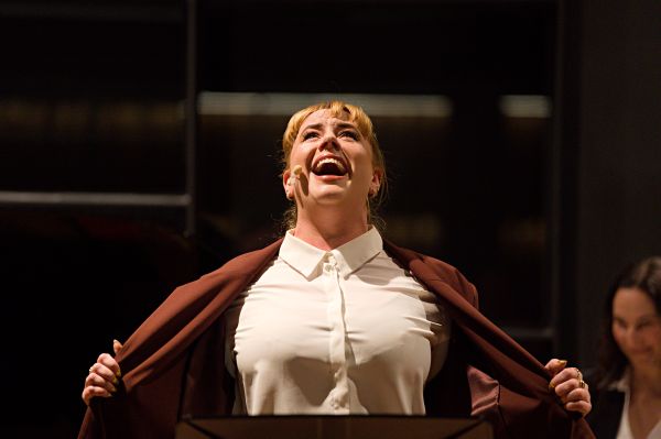 A person wearing a white button-up shirt and a brown jacket is standing indoors, holding the jacket open with both hands. The background is dark with blurred architectural details and lighting.