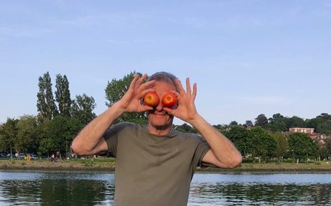 A man wearing a T-shirt standing outside in front of a river and greenery, holding two apples up in front of his eyes