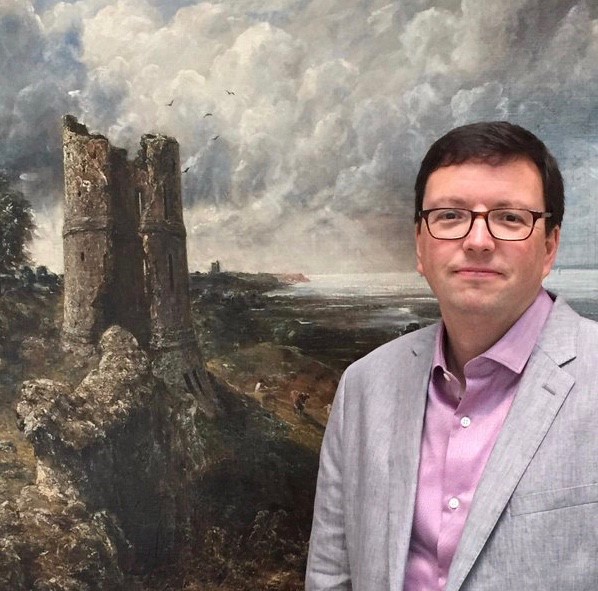A man wearing glasses and a light grey blazer over a purple shirt stands in front of a large, dramatic painting featuring a stormy sky and the ruins of a tower by a rocky shoreline. The scene behind him appears to depict a coastal landscape with dark clouds looming overhead.