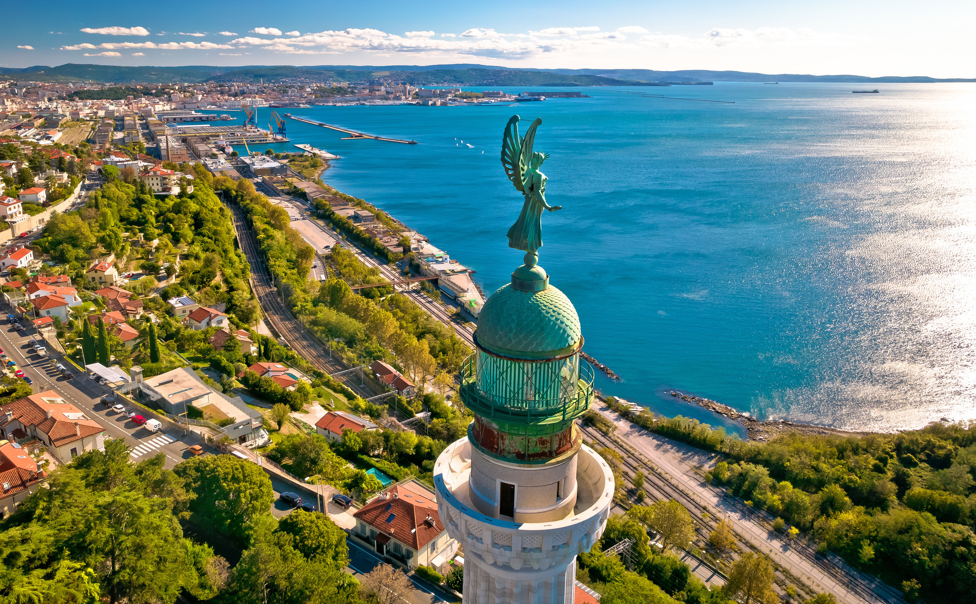 A view across a city, looking out to sea, with mountains in the background and a lighthouse in the foreground.