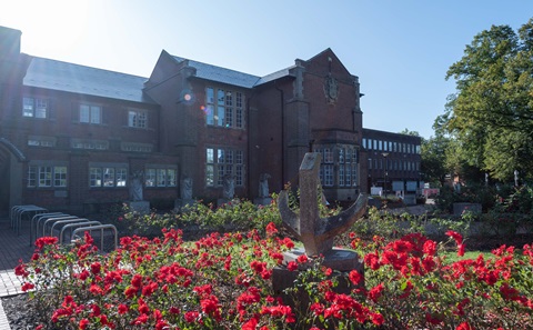 A large red brick building under a blue sky, with a large flowerbed with red flowers in the foreground