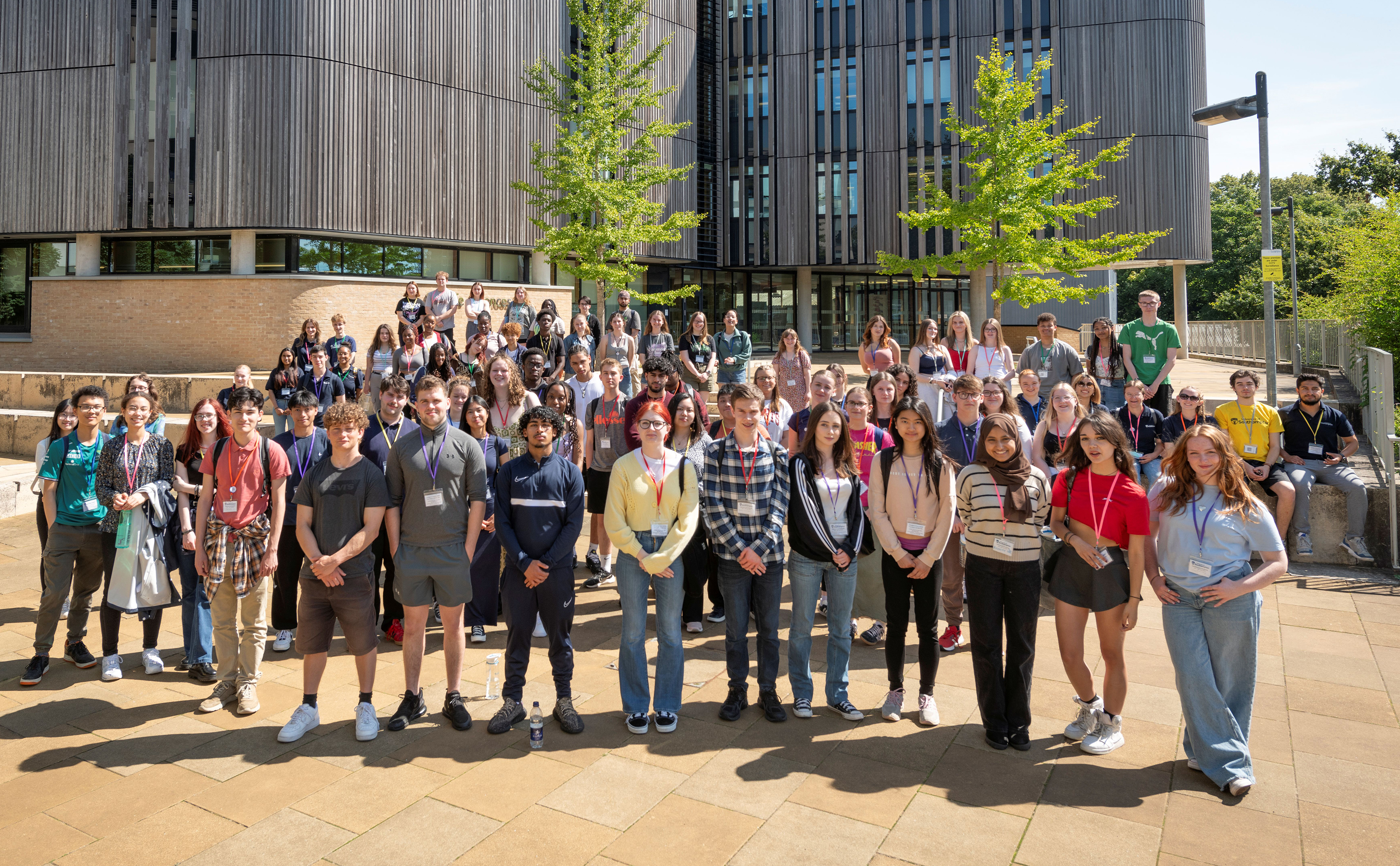 A large, posed, group of young people standing outside a large wood-clad building on a university campus.
