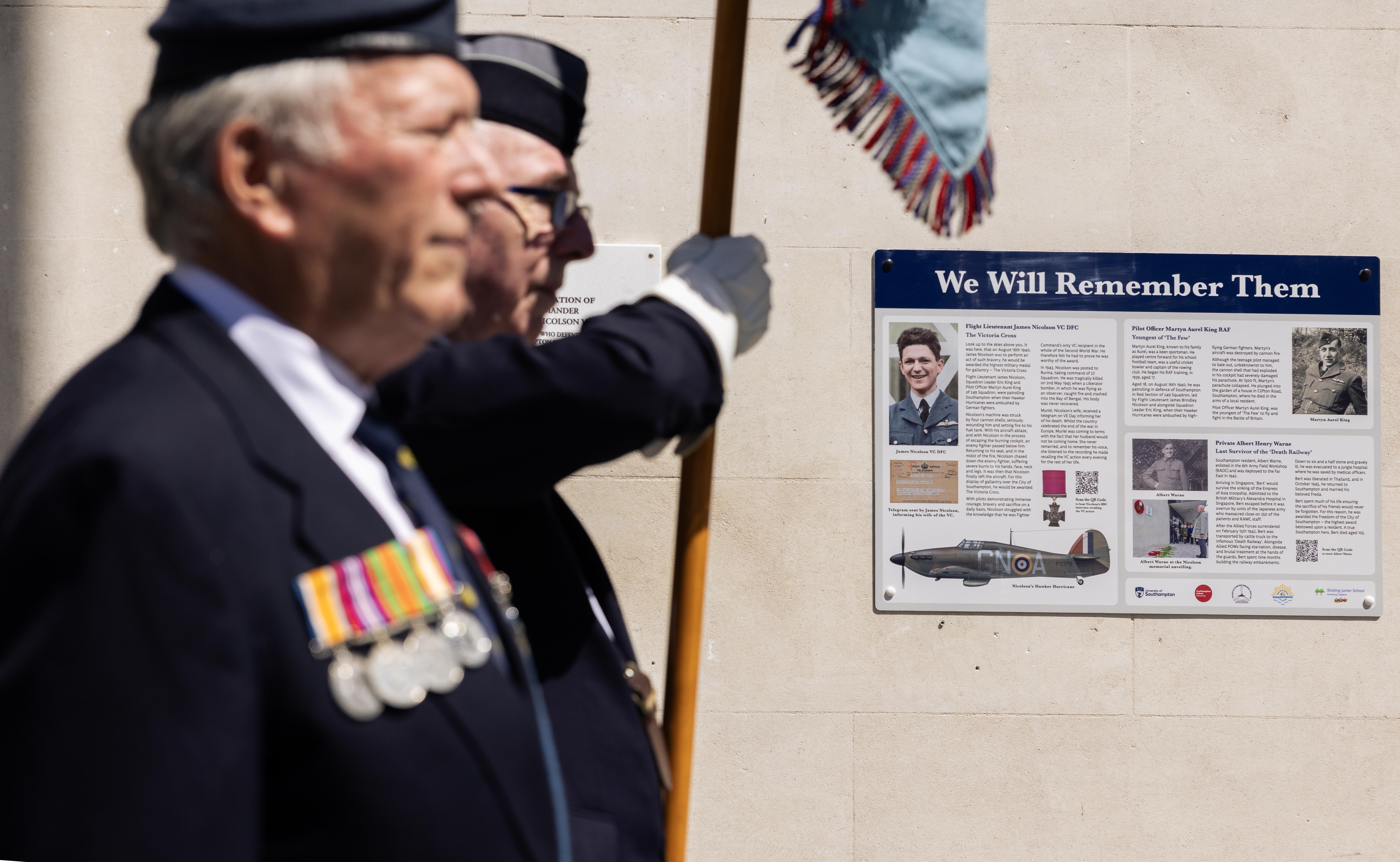 Two smartly dressed men stand to attention in front of an information board 