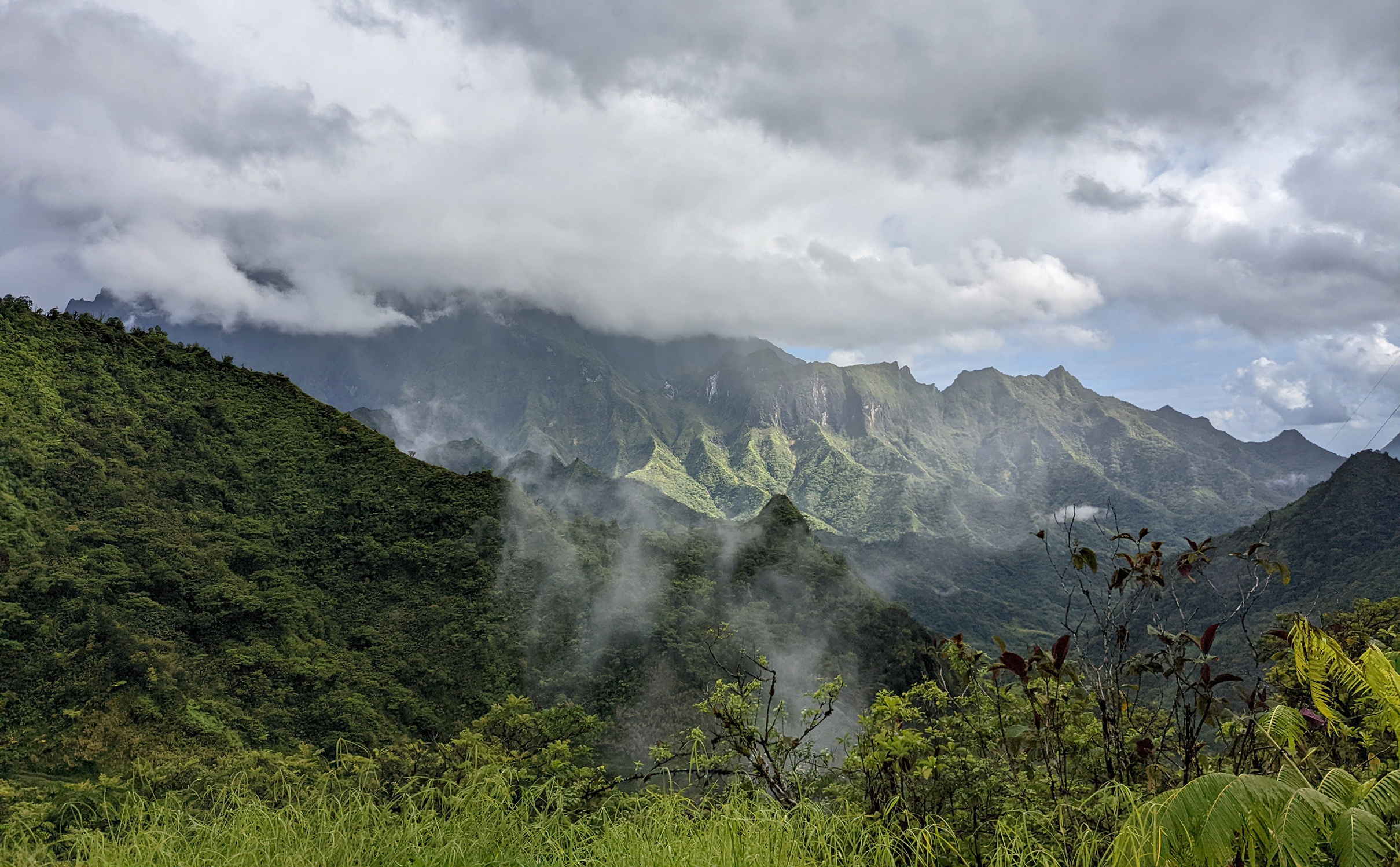 A lush mountain landscape with green hills and mist under a cloudy sky.