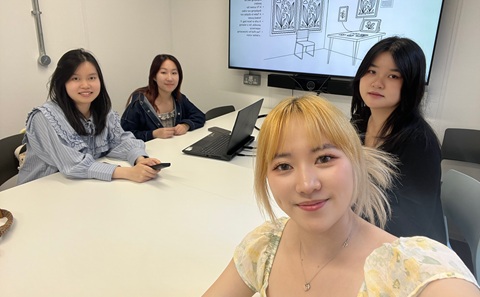 Four women sat around a white table with a laptop on it, smiling at the camera. 