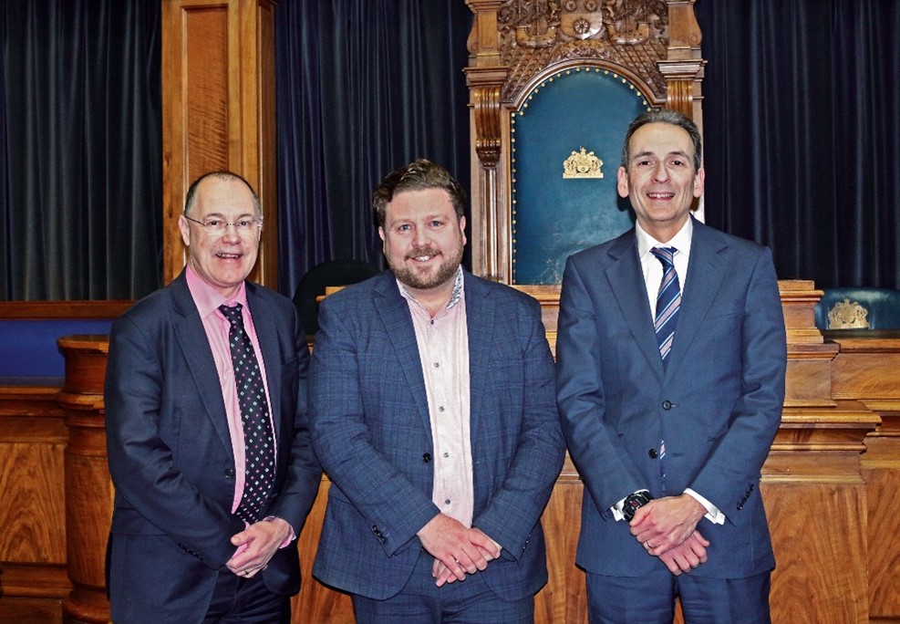 Three people are standing and smiling for an official-looking photograph. In the background is an area of wood panelling and dark velvet curtains. 