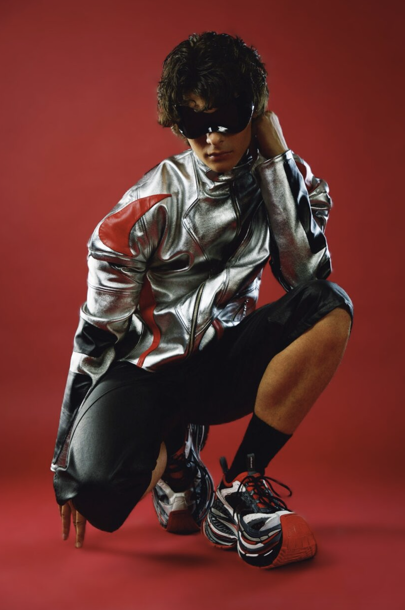A person poses against a red background wearing a shiny silver jacket with red accents, black shorts, black socks, and sneakers. They are crouching with one hand on the ground and wearing dark sunglasses.