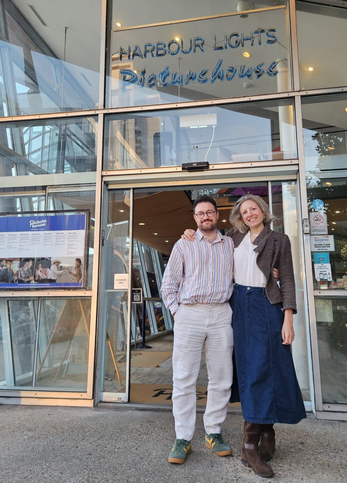 Two people stand close together in front of the glass entrance to Harbour Lights Picturehouse, with one person’s arm around the other’s shoulder. A poster is visible to the left of the door, and the interior lobby with more posters can be seen through the glass.