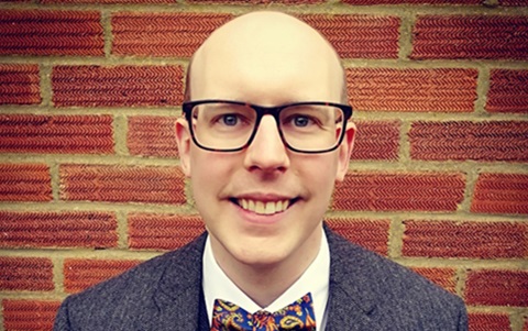 Man wearing glasses, a blazer, waistcoat, white shirt and bow tie, smiling at the camera. He is in front of a brick wall.