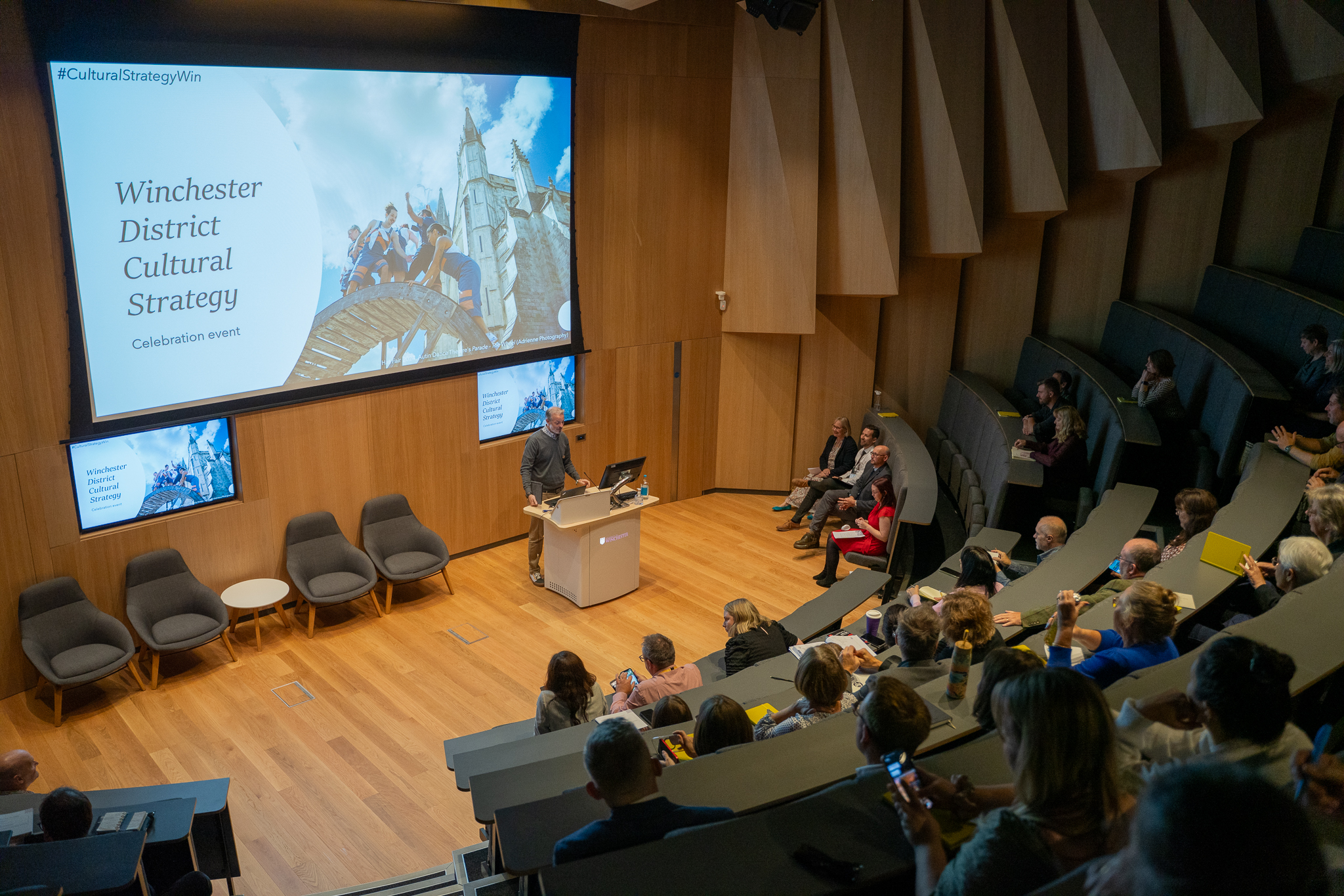 Audience members sit in a modern lecture hall, listening to a speaker at a podium during the "Winchester District Cultural Strategy" celebration event, with a large presentation screen displaying event details. Some attendees are taking notes or photos as they engage with the presentation.