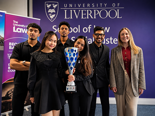 A group of six people pose in front of a purple University of Liverpool School of Law and Social Justice backdrop, with two students at the centre holding a large silver-and-blue trophy. Five of the group are dressed mostly in black smart-casual clothing, while one woman on the right wears a checked blazer, red top and beige trousers.