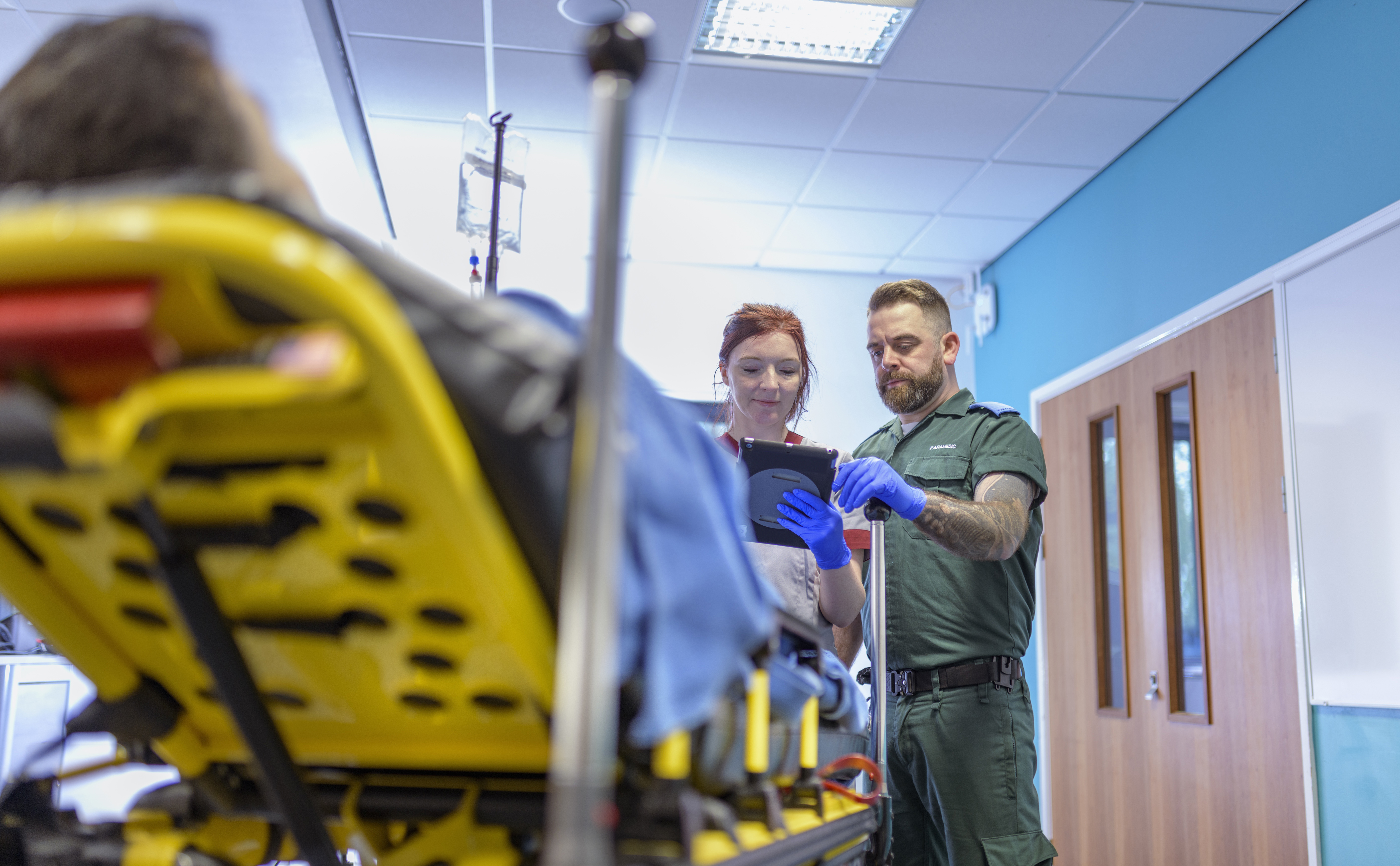 A nurse and a paramedic stood looking at a clipboard, facing a patient on an ambulance trolley who is seen from behind.