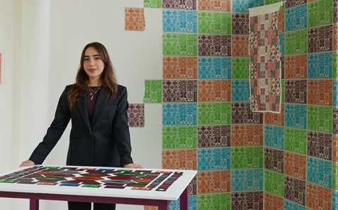 Woman with long dark hair wearing a black blazer stood behind a table with a pattern of tiles on it. Behind her and to the right is a white wall with a printed tile design on it.