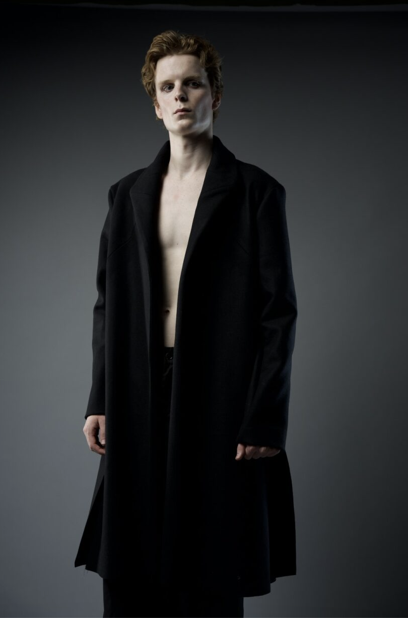 A person with short, curly hair stands against a dark gray background wearing an open black coat that reveals a bare chest. The lighting emphasizes the contrast between their pale skin and dark clothing.