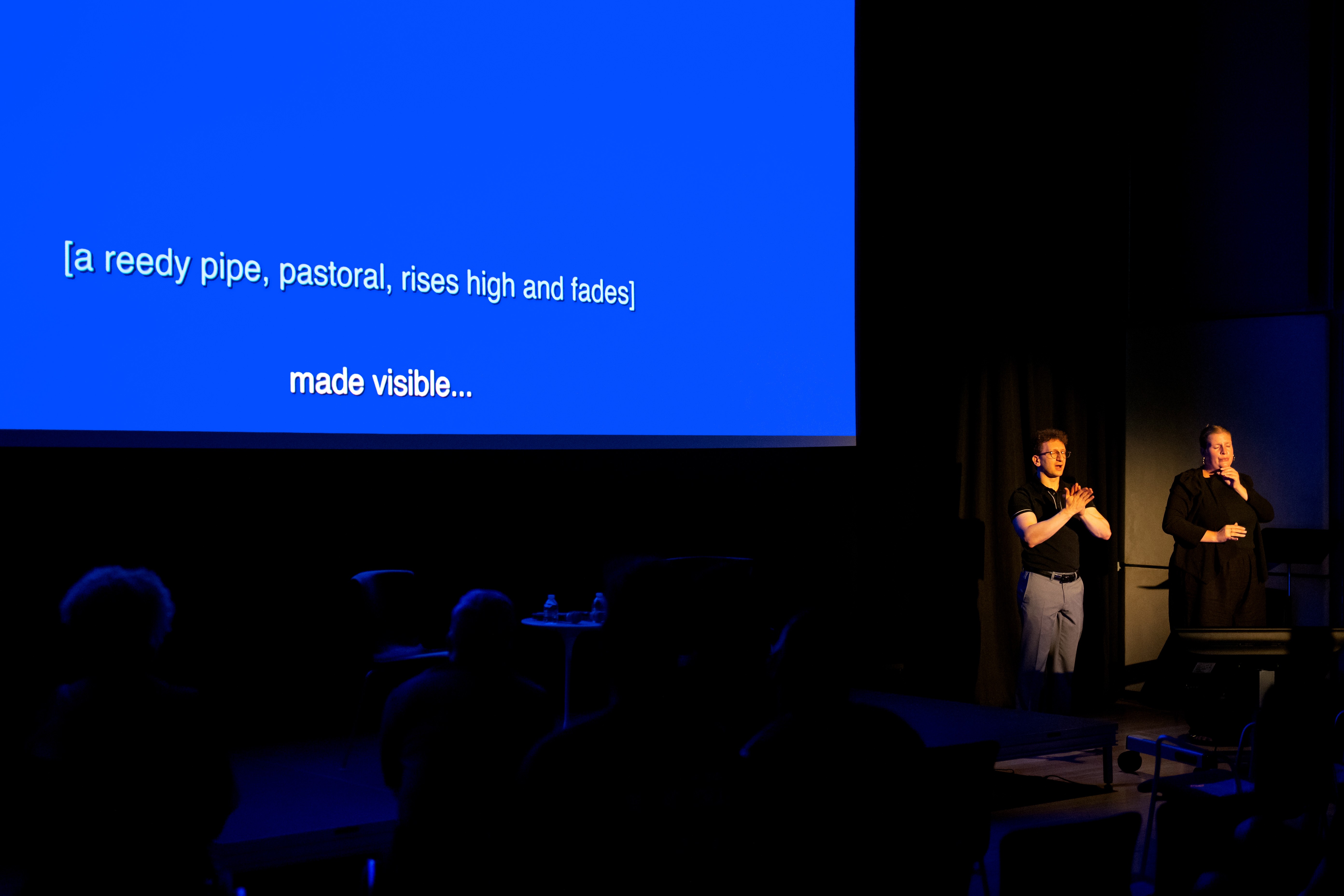 The image shows a darkened presentation room with a large blue screen displaying white text that reads: 