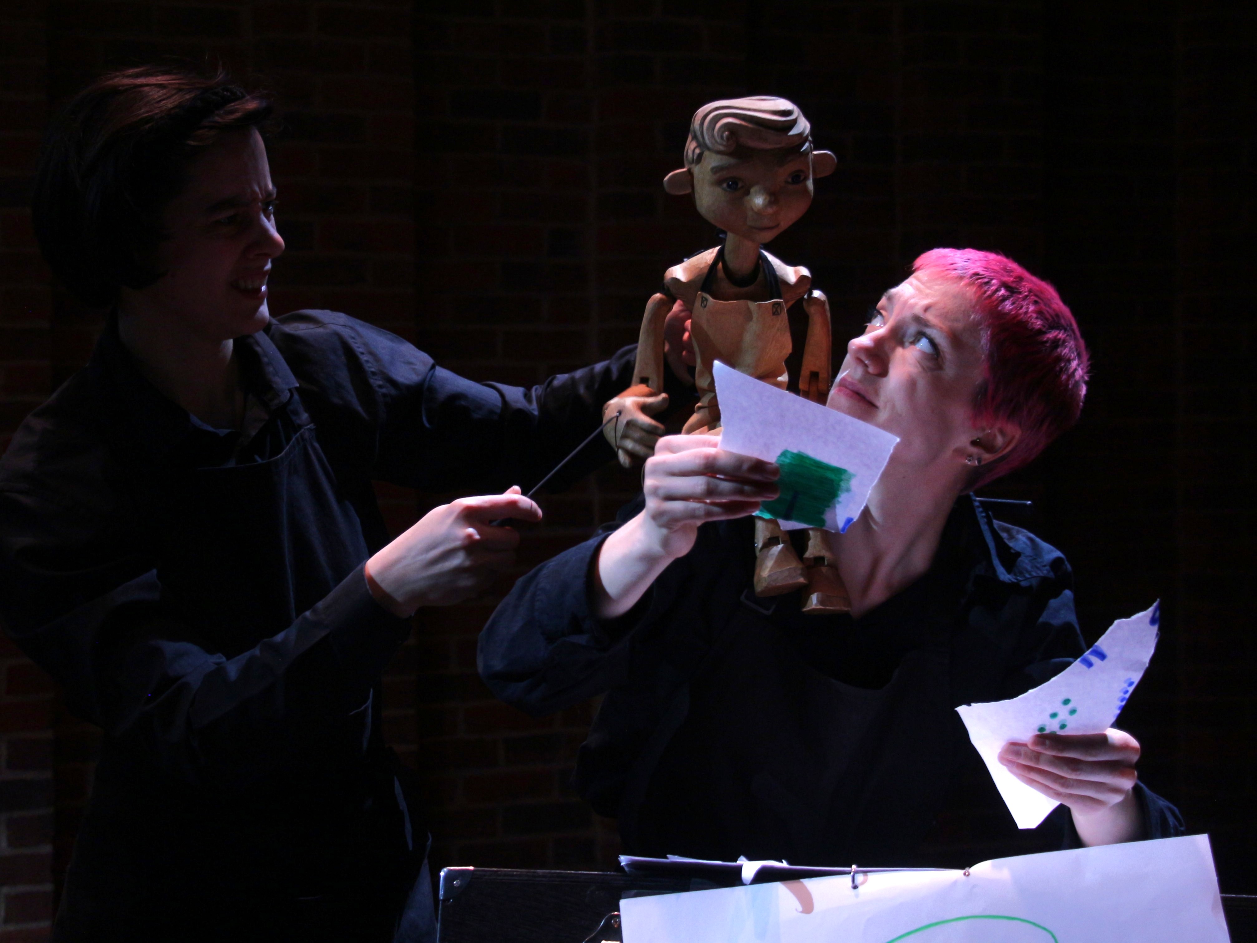 Two performers dressed in black manipulate a wooden puppet seated on a table covered with a star-patterned cloth, while one holds a container of colored pencils. The scene is lit dramatically against a dark background, suggesting a stage performance.