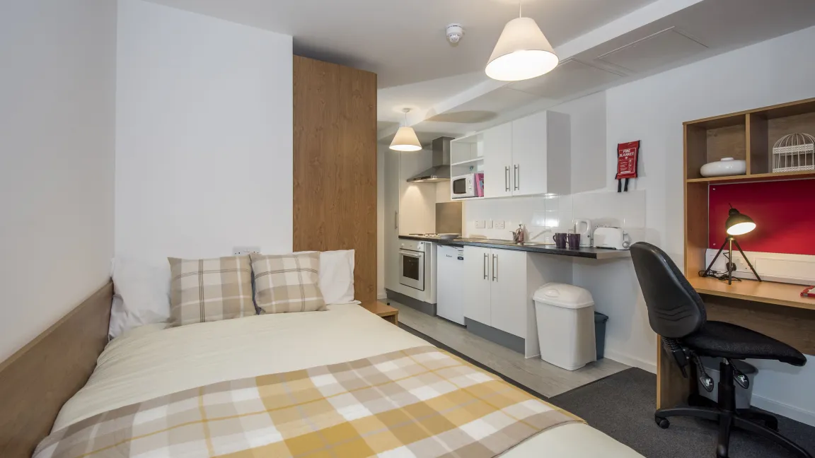 A modern studio flat showing bed, desk and kitchenette.