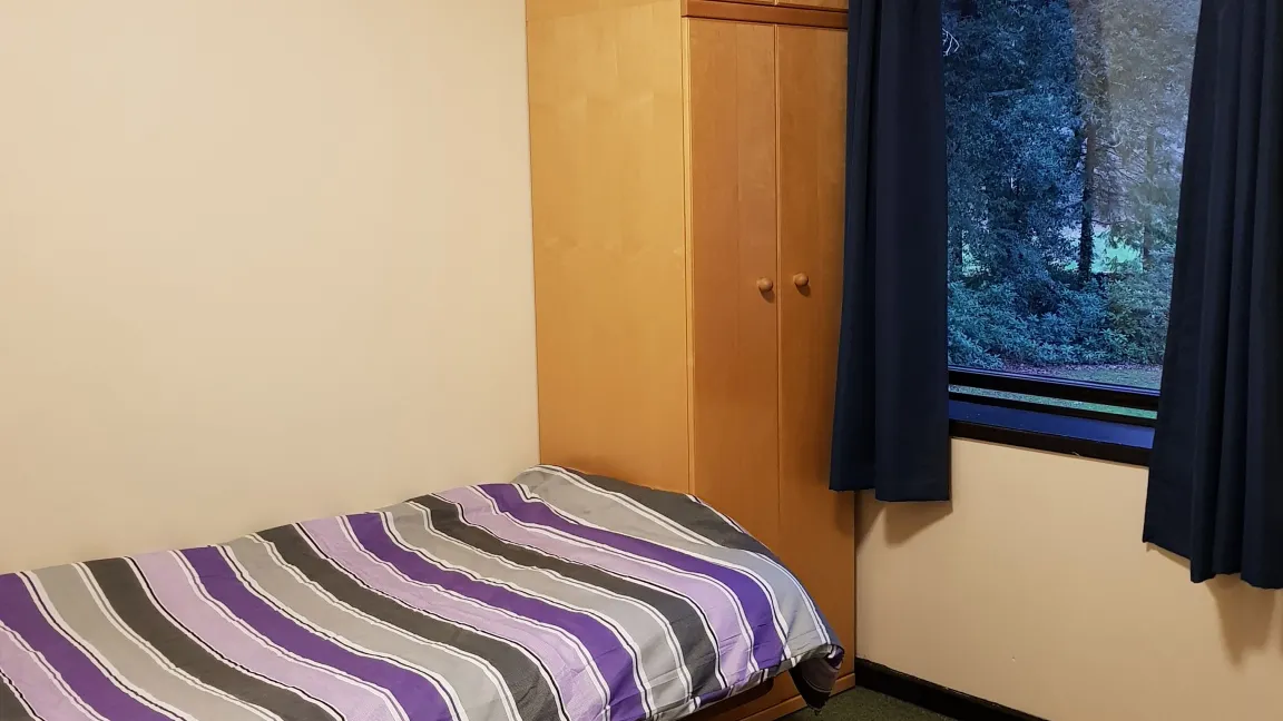 A student bedroom showing a single bed with stripey covers, a tall wooden wardrobe and a window looking out to a landscape filled with trees.