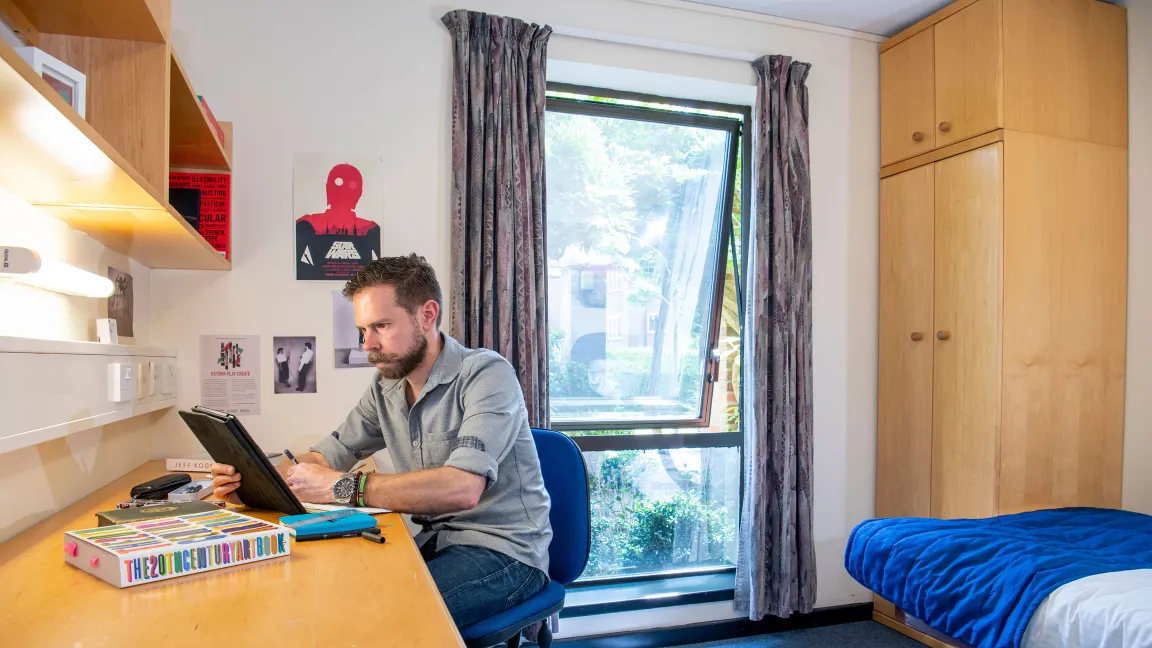 A student sits looking at a tablet computer while working at a desk. A bed and wardrobe can be seen behind him, an open window lets light into the room.