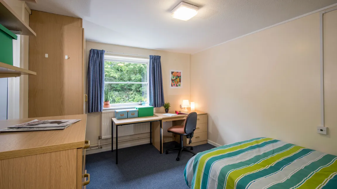 A large student bedroom. The room contains a double bed with striped covers, a large desk, a wooden wardrobe and chest of drawers.