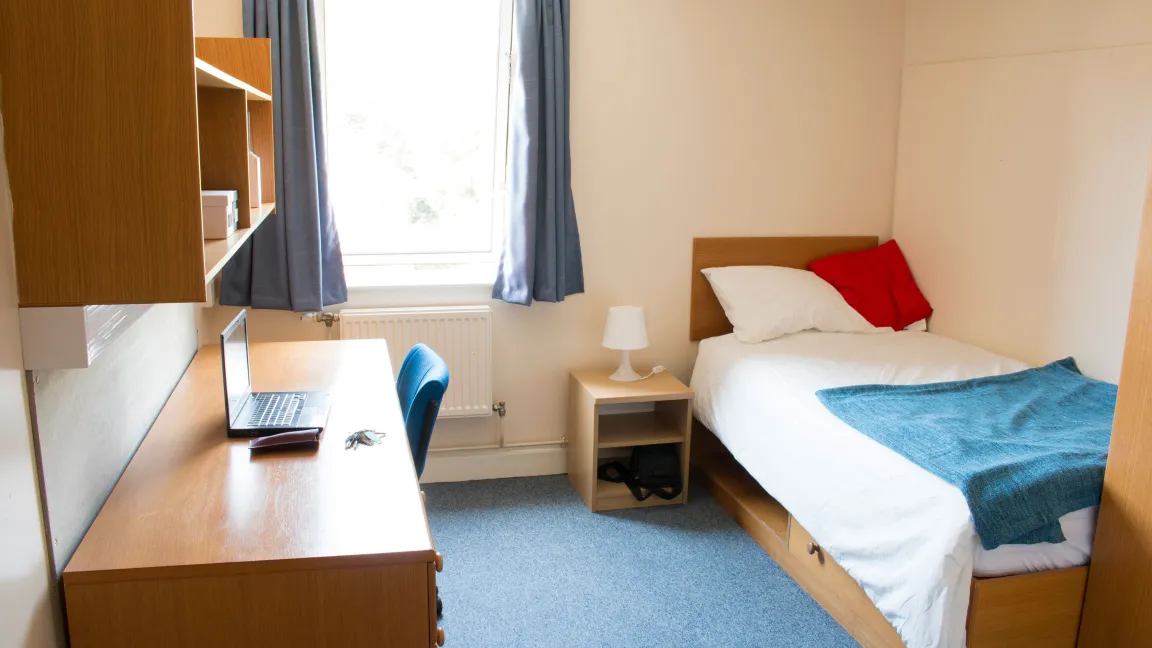 A student bedroom. A section of blue carpet separates a single bed with patterned sheets and a large wooden desk. A window on the rear wall lets light in.